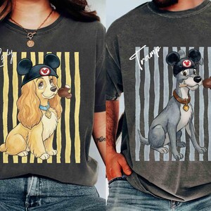 May include: Two charcoal gray t-shirts featuring Disney characters. The left shirt shows Lady with the text Lady, wearing mouse ears and holding a treat. The right shirt shows Tramp with the text Tramp, wearing mouse ears and holding a treat, both set against vertical stripes.