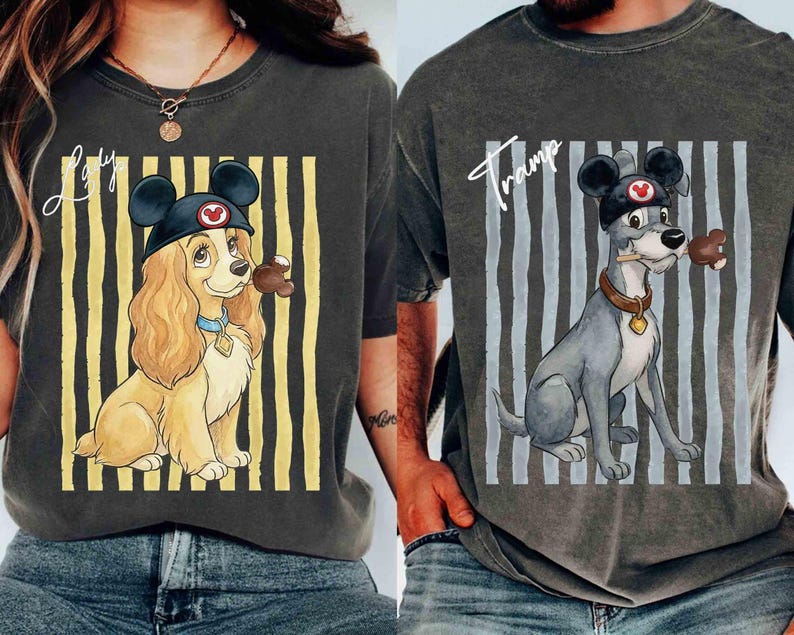 May include: Two charcoal gray t-shirts featuring Disney characters. The left shirt shows Lady with the text Lady, wearing mouse ears and holding a treat. The right shirt shows Tramp with the text Tramp, wearing mouse ears and holding a treat, both set against vertical stripes.