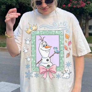 May include: A cream-colored t-shirt featuring a graphic print of Olaf the snowman and Bruni the salamander inside a green ornate frame. The design includes a pink bow, a gold crown, small snowmen, colorful autumn leaves, and blue snowflakes.
