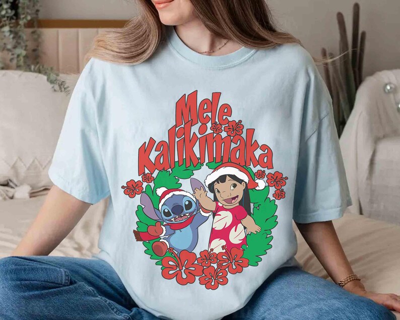 May include: Light blue t-shirt featuring the text "Mele Kalikimaka" in red, with a cartoon illustration of Stitch and Lilo in Santa hats, surrounded by red flowers and green leaves. The shirt is a relaxed fit.