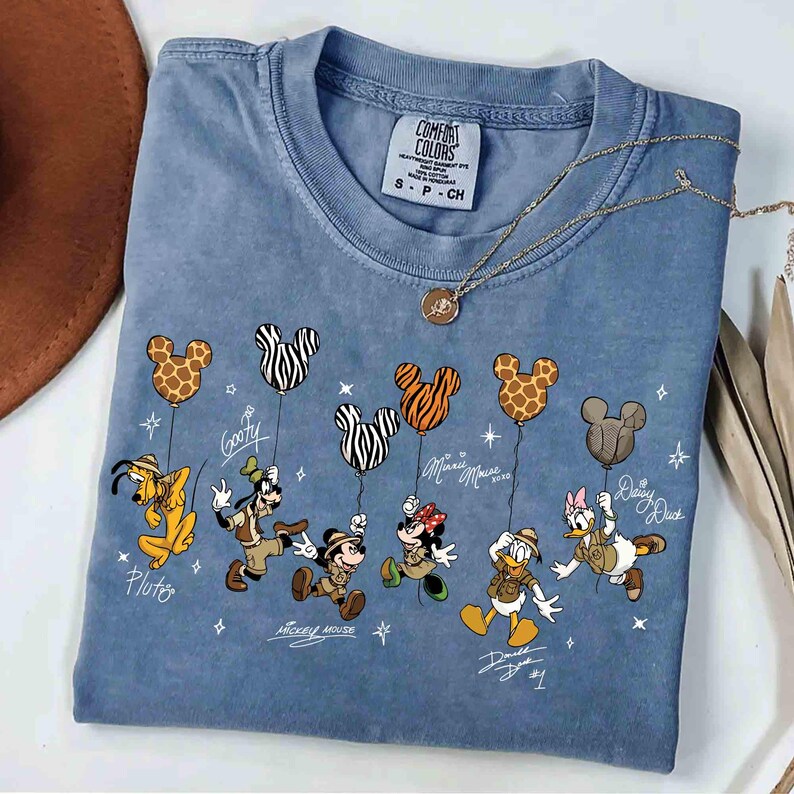 May include: A blue Comfort Colors t-shirt featuring Disney characters holding Mickey Mouse-shaped balloons. The characters include Pluto, Goofy, Mickey Mouse, Minnie Mouse, Donald Duck, and Daisy Duck. A gold necklace is draped across the shirt.