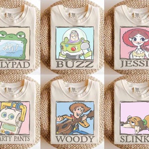 May include: Six cream-colored t-shirts, each featuring a different cartoon character design. The designs include a frog, Buzz Lightyear, Jessie, Smarty Pants, Woody, and Slinky. Each design is framed with a colored border and character name.