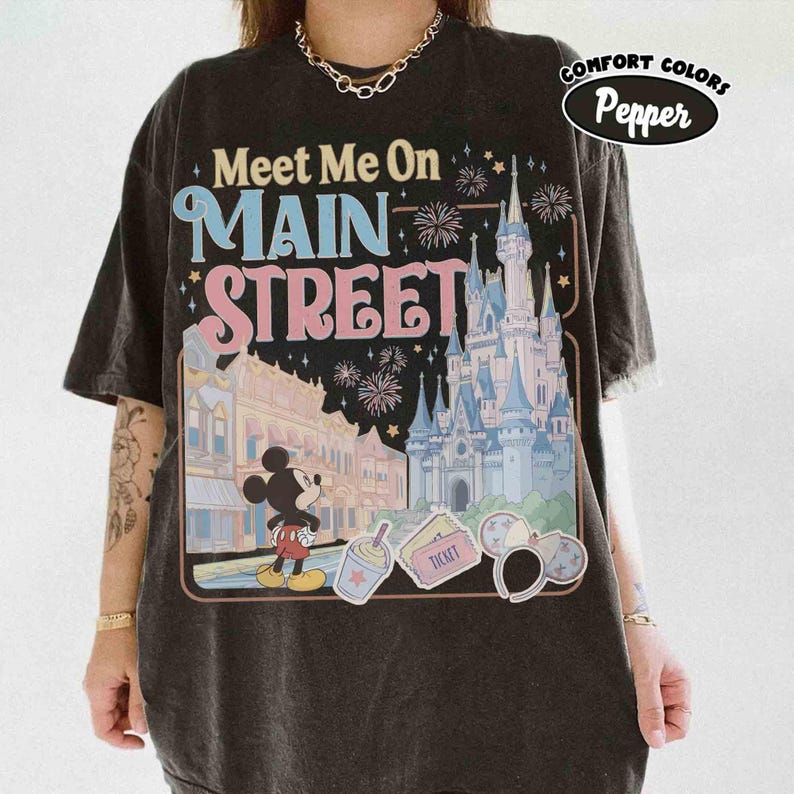May include: Black t-shirt with a graphic featuring the text "Meet Me On Main Street" above an illustration of a castle, Mickey Mouse, and other park-themed icons. The shirt has a vintage, distressed look.