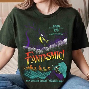 K&ouml;nnte beinhalten: Dunkelgr&uuml;nes T-Shirt mit dem Wort "FANTASMIC!" in Gelb und Orange und einer Grafik eines Drachen. Der Text auf dem Shirt lautet "GOOD CLASHES WITH EVIL IN A NIGHTTIME SPECTACULAR" und "RIVERS OF AMERICA NEW ORLEANS SQUARE FRONTIERLAND".