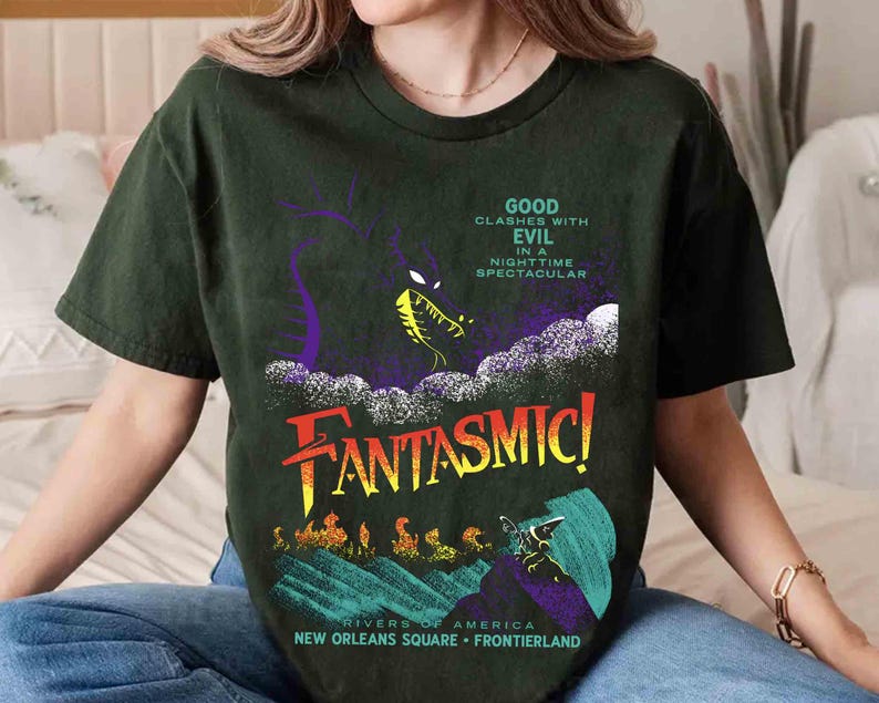 K&ouml;nnte beinhalten: Dunkelgr&uuml;nes T-Shirt mit dem Wort "FANTASMIC!" in Gelb und Orange und einer Grafik eines Drachen. Der Text auf dem Shirt lautet "GOOD CLASHES WITH EVIL IN A NIGHTTIME SPECTACULAR" und "RIVERS OF AMERICA NEW ORLEANS SQUARE FRONTIERLAND".