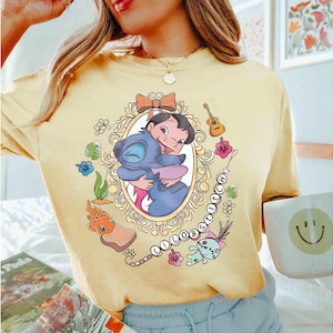 May include: Yellow t-shirt featuring a framed graphic of Lilo and Stitch hugging. The design includes a bow, guitar, flowers, and the word "Lilo & Stitch" in a chain. The shirt is a light yellow color.