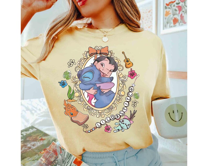 May include: Yellow t-shirt featuring a framed graphic of Lilo and Stitch hugging. The design includes a bow, guitar, flowers, and the word "Lilo & Stitch" in a chain. The shirt is a light yellow color.