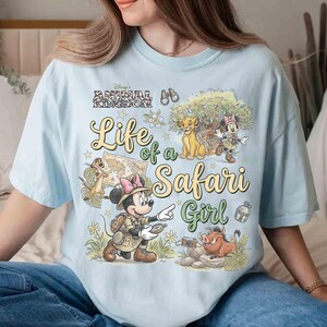May include: Light blue t-shirt with the text "Life of a Safari Girl" and Disney's Animal Kingdom logo. The graphic features cartoon characters including Simba, Timon, Pumbaa, and Minnie Mouse in safari attire.