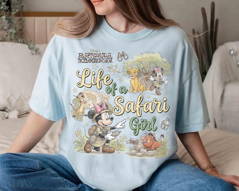 May include: Light blue t-shirt with the text "Life of a Safari Girl" and Disney's Animal Kingdom logo. The graphic features cartoon characters including Simba, Timon, Pumbaa, and Minnie Mouse in safari attire.