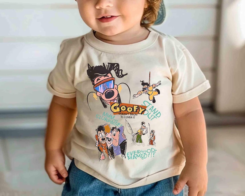 May include: A toddler wearing a beige t-shirt with a cartoon graphic featuring Goofy and the text "Goofy Doesn't Begin To Cover It", "Stand Out", "Mmm Slimerace", and "Everybody Mambo!!"