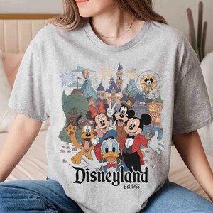 May include: A gray t-shirt featuring a colorful graphic of Disneyland characters, including Mickey Mouse, Minnie Mouse, Donald Duck, and Goofy. The Disneyland logo and "Est. 1955" are printed below the characters. The shirt has a crew neck.