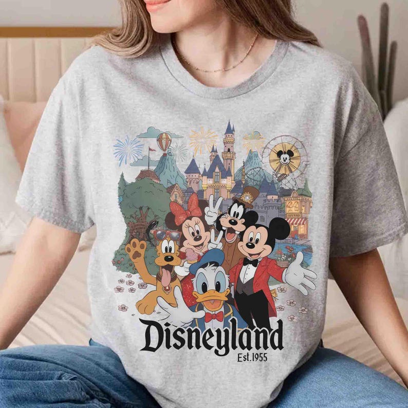 May include: A gray t-shirt featuring a colorful graphic of Disneyland characters, including Mickey Mouse, Minnie Mouse, Donald Duck, and Goofy. The Disneyland logo and "Est. 1955" are printed below the characters. The shirt has a crew neck.