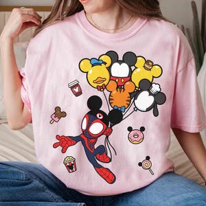 Puede incluir: Camiseta rosa claro con una ilustraci&oacute;n de dibujos animados de un personaje similar a Spider-Man sosteniendo globos con forma de Mickey Mouse. Tambi&eacute;n hay iconos de comida de dibujos animados, como una taza de caf&eacute; y helado.