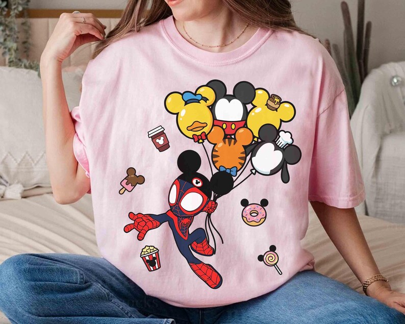 Puede incluir: Camiseta rosa claro con una ilustraci&oacute;n de dibujos animados de un personaje similar a Spider-Man sosteniendo globos con forma de Mickey Mouse. Tambi&eacute;n hay iconos de comida de dibujos animados, como una taza de caf&eacute; y helado.