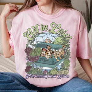 May include: Pink t-shirt with a graphic featuring Disney characters on a boat ride with the text "Get in Lose" and "We're learning about Lettuce". The design includes green and purple botanical elements.
