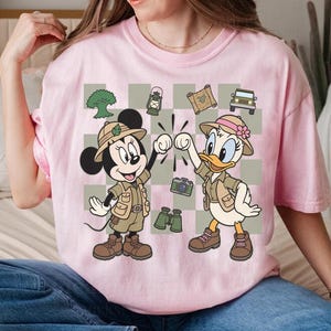 May include: Pink t-shirt featuring Mickey Mouse and Daisy Duck in safari outfits, giving a high-five. The design includes a checkered background with safari-themed icons like a jeep, camera, and binoculars. The shirt is a light pink color.