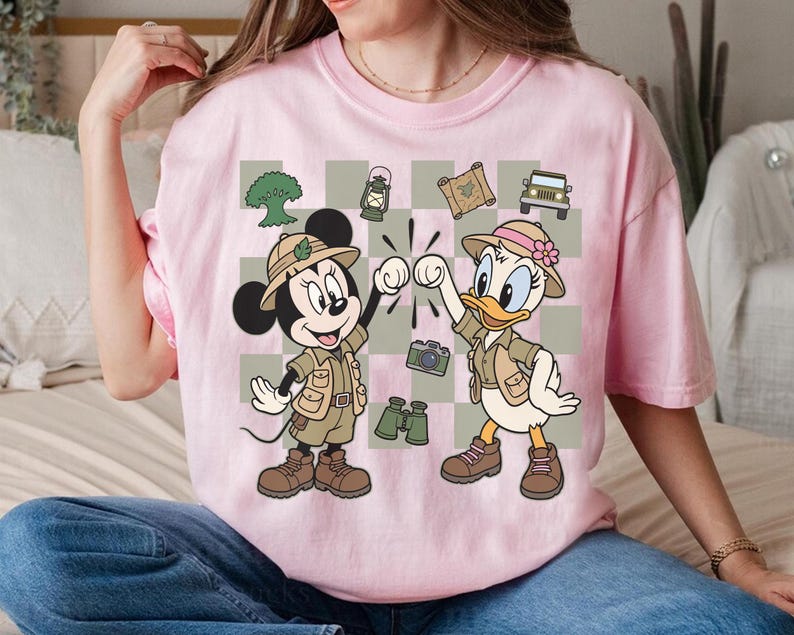 May include: Pink t-shirt featuring Mickey Mouse and Daisy Duck in safari outfits, giving a high-five. The design includes a checkered background with safari-themed icons like a jeep, camera, and binoculars. The shirt is a light pink color.