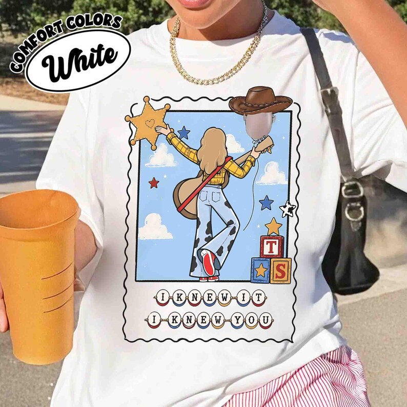 May include: A white t-shirt featuring a graphic print of a person with long hair wearing a yellow plaid shirt and cow-print pants, holding a guitar. The design includes a sheriff star, a cowboy hat, building blocks, and the text I KNEW IT I KNEW YOU.