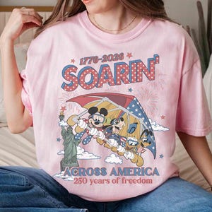 May include: Pink t-shirt featuring Mickey Mouse, Goofy, and Donald Duck soaring in a hang glider with the Statue of Liberty. The shirt has the text "SOARIN'" and "ACROSS AMERICA 250 years of freedom" in a patriotic design.