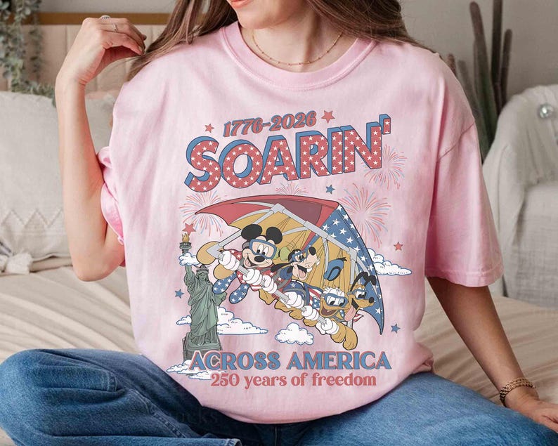 May include: Pink t-shirt featuring Mickey Mouse, Goofy, and Donald Duck soaring in a hang glider with the Statue of Liberty. The shirt has the text "SOARIN'" and "ACROSS AMERICA 250 years of freedom" in a patriotic design.
