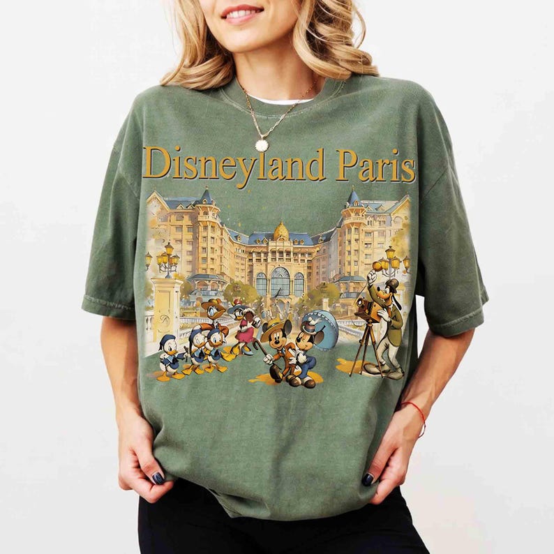 May include: Olive green t-shirt featuring the text "Disneyland Paris" above a colorful illustration of Disney characters in front of a grand building. The graphic includes Mickey Mouse, Donald Duck, and Goofy.
