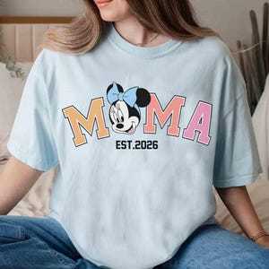 May include: Light blue t-shirt featuring the word "MOMA" in orange and pink block letters, with a Minnie Mouse graphic. The text "EST. 2026" is below the graphic. The shirt is worn by a person.