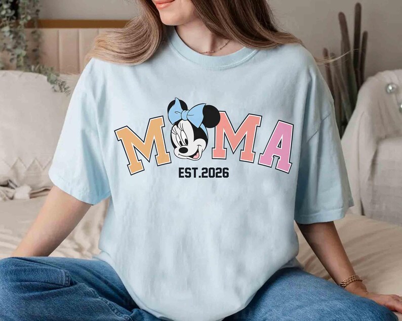 May include: Light blue t-shirt featuring the word "MOMA" in orange and pink block letters, with a Minnie Mouse graphic. The text "EST. 2026" is below the graphic. The shirt is worn by a person.