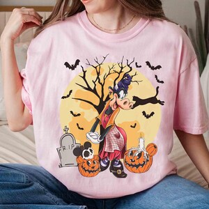 Peut inclure: T-shirt rose clair avec un motif d'Halloween. Le motif comprend un personnage de dessin anim&eacute; d&eacute;guis&eacute; en sorci&egrave;re, des chauves-souris, des citrouilles, une pierre tombale et une pleine lune. Le t-shirt est de style d&eacute;contract&eacute; &agrave; manches courtes.