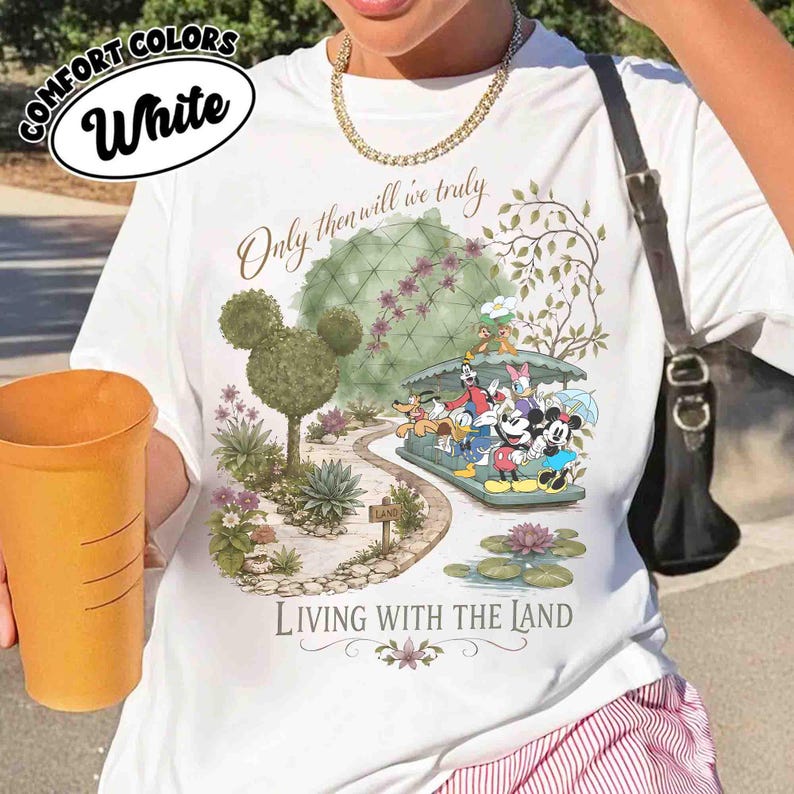 May include: White t-shirt with a colorful graphic featuring Mickey Mouse and friends on a boat ride. The shirt has the text "Only then will we truly Living with the Land" and "Comfort Colors White" in a circular design.