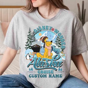 May include: A gray t-shirt featuring a Disney-themed design. The graphic includes Pluto in winter attire, the words "Disney Wonder Alaska Cruise", and a space for a custom name. The design incorporates winter elements like snow, trees, and a cruise ship.