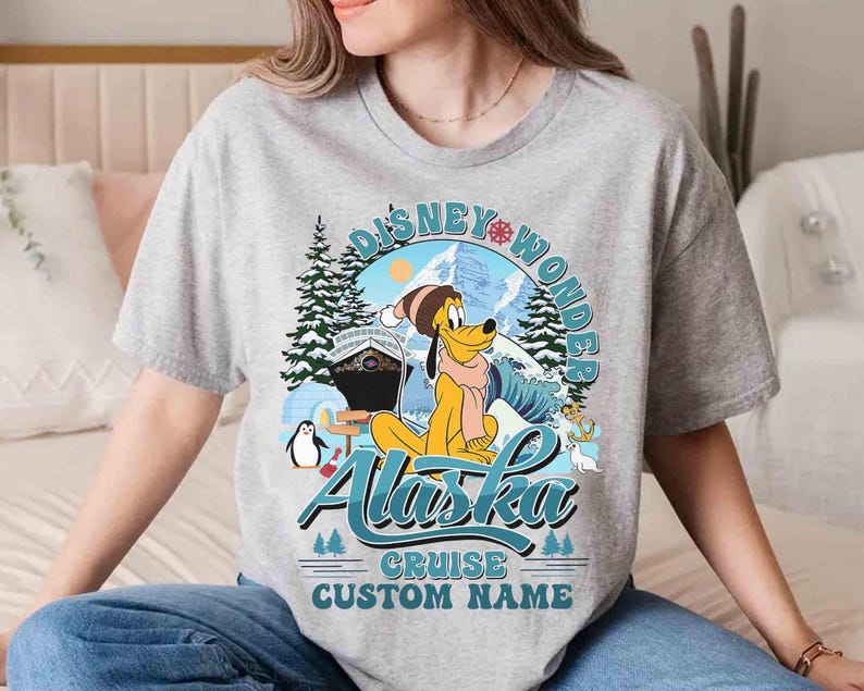 May include: A gray t-shirt featuring a Disney-themed design. The graphic includes Pluto in winter attire, the words "Disney Wonder Alaska Cruise", and a space for a custom name. The design incorporates winter elements like snow, trees, and a cruise ship.