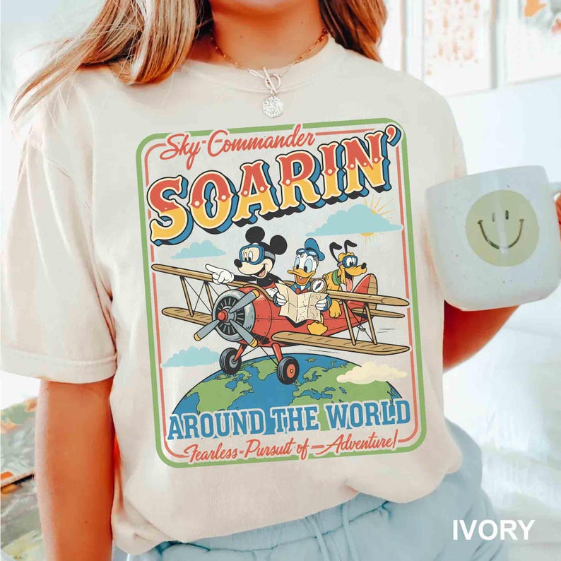 May include: Ivory-colored t-shirt featuring a vintage-style graphic of Mickey Mouse, Donald Duck, and Pluto in a red biplane. The graphic reads "Sky Commander Soarin' Around the World". A white mug with a smiley face is visible.