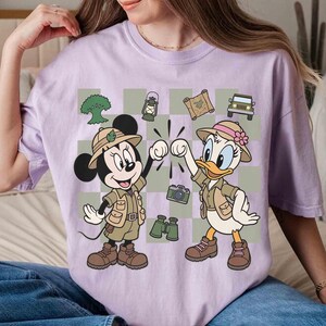 May include: A light purple t-shirt featuring a cartoon graphic of Mickey Mouse and Daisy Duck dressed in safari outfits. The characters are fist-bumping against a checkered background with safari-themed icons like a jeep and binoculars.