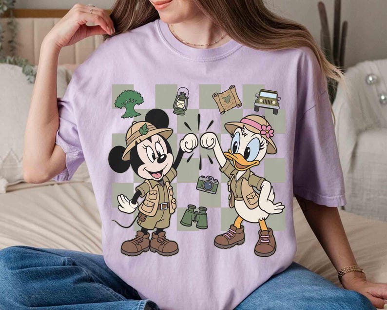 May include: A light purple t-shirt featuring a cartoon graphic of Mickey Mouse and Daisy Duck dressed in safari outfits. The characters are fist-bumping against a checkered background with safari-themed icons like a jeep and binoculars.