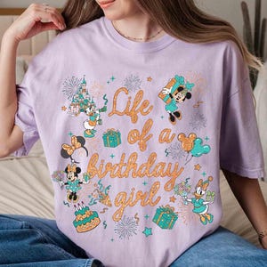 May include: A light purple t-shirt with the text "Life of a birthday girl" in orange script. The shirt features cartoon images of Minnie Mouse, Daisy Duck, and other Disney characters, along with fireworks, gifts, and a birthday cake.