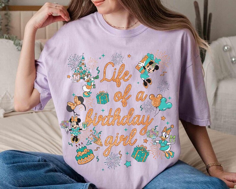 May include: A light purple t-shirt with the text "Life of a birthday girl" in orange script. The shirt features cartoon images of Minnie Mouse, Daisy Duck, and other Disney characters, along with fireworks, gifts, and a birthday cake.