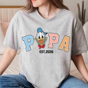 May include: A gray t-shirt featuring a cartoon image of Donald Duck with the word "POPA" in blue, pink, and orange block letters. The text "EST.2026" is below the word "POPA".