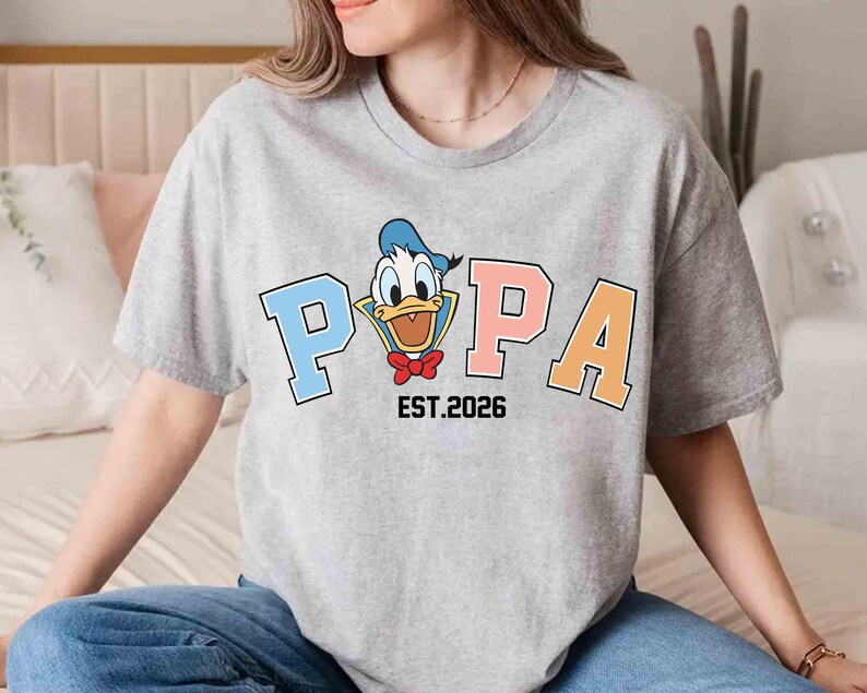 May include: A gray t-shirt featuring a cartoon image of Donald Duck with the word "POPA" in blue, pink, and orange block letters. The text "EST.2026" is below the word "POPA".