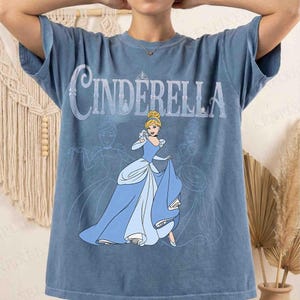 Op de afbeelding: Blauw T-shirt met het woord "Cinderella" in een decoratief lettertype. Het shirt heeft een afbeelding van Assepoester in haar baljurk. Het ontwerp bevat twee extra vage Assepoester-omtrekken. Het shirt heeft een gedempte blauwe kleur.