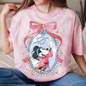 May include: A pale pink t-shirt with a cartoon Mickey Mouse design. The image shows Mickey giving a thumbs up, framed by a pink bow and ribbon. Text reads "Some imagination huh?" within an oval.