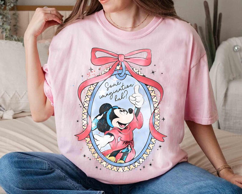 May include: A pale pink t-shirt with a cartoon Mickey Mouse design. The image shows Mickey giving a thumbs up, framed by a pink bow and ribbon. Text reads "Some imagination huh?" within an oval.