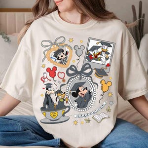 Puede incluir: Camiseta color crema con personajes de Disney con atuendo de graduaci&oacute;n. Mickey Mouse, el Pato Donald y Goofy est&aacute;n representados con gorras y togas de graduaci&oacute;n, rodeados de elementos decorativos como globos y un marco en forma de coraz&oacute;n.