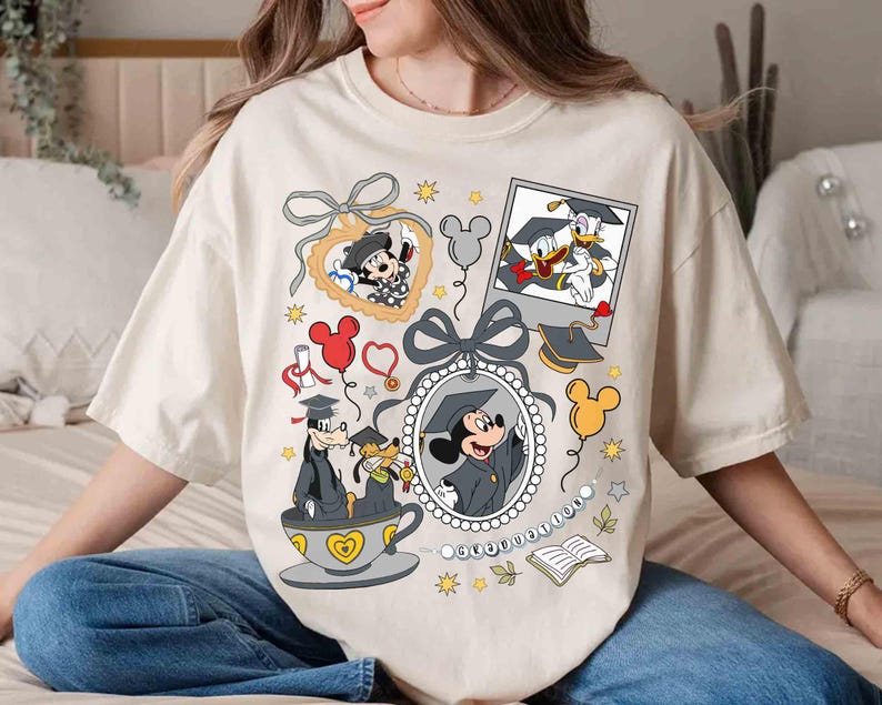 Puede incluir: Camiseta color crema con personajes de Disney con atuendo de graduaci&oacute;n. Mickey Mouse, el Pato Donald y Goofy est&aacute;n representados con gorras y togas de graduaci&oacute;n, rodeados de elementos decorativos como globos y un marco en forma de coraz&oacute;n.