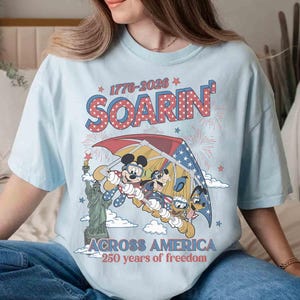 May include: Light blue t-shirt featuring a graphic of Mickey Mouse, Donald Duck, and Goofy soaring over the Statue of Liberty. The design includes the text "1776-2026 Soarin' Across America 250 years of freedom" in red, white, and blue.