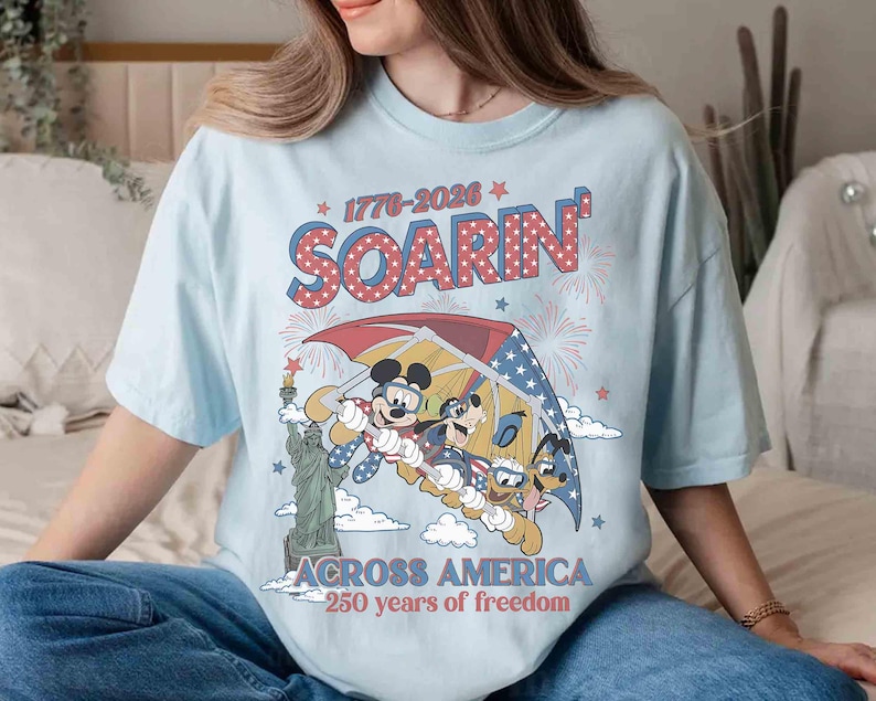 May include: Light blue t-shirt featuring a graphic of Mickey Mouse, Donald Duck, and Goofy soaring over the Statue of Liberty. The design includes the text "1776-2026 Soarin' Across America 250 years of freedom" in red, white, and blue.