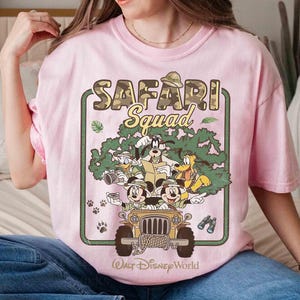 May include: A pale pink t-shirt with a graphic of Disney characters in a safari jeep. The text "SAFARI Squad" is above the characters, with "Walt Disney World" below. The design includes Mickey Mouse, Goofy, Donald Duck, and Pluto, in a jungle setting.