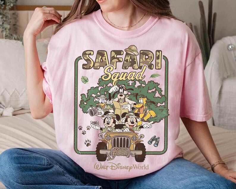 May include: A pale pink t-shirt with a graphic of Disney characters in a safari jeep. The text "SAFARI Squad" is above the characters, with "Walt Disney World" below. The design includes Mickey Mouse, Goofy, Donald Duck, and Pluto, in a jungle setting.