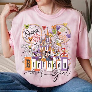 May include: Pink t-shirt featuring a colorful graphic of a castle, balloons, and various cartoon characters. The shirt has the words "Name" and "Birthday Girl" printed on it. The design includes fireworks and a spider web.