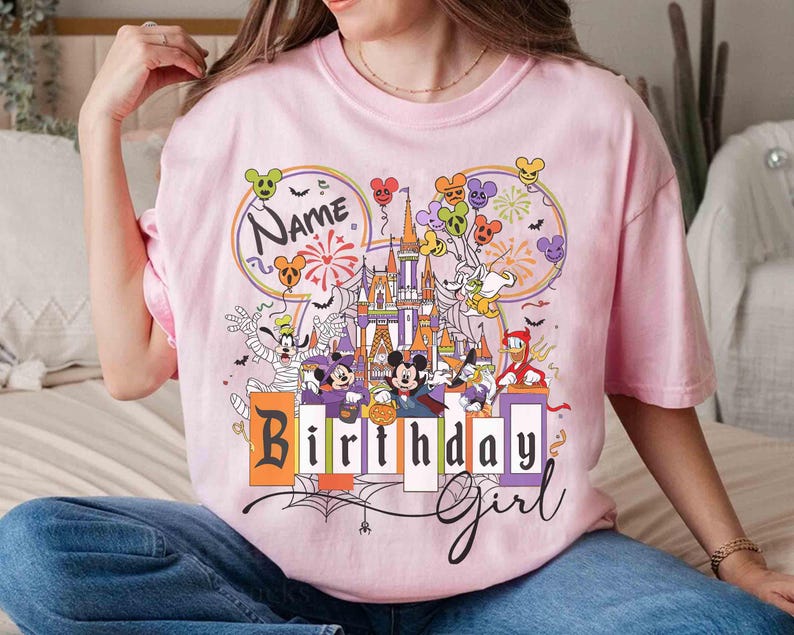 May include: Pink t-shirt featuring a colorful graphic of a castle, balloons, and various cartoon characters. The shirt has the words "Name" and "Birthday Girl" printed on it. The design includes fireworks and a spider web.