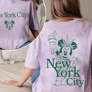 May include: A light purple t-shirt with the text "New York City" in green, along with a green graphic of Minnie Mouse as the Statue of Liberty. The back of the shirt also features the same design.